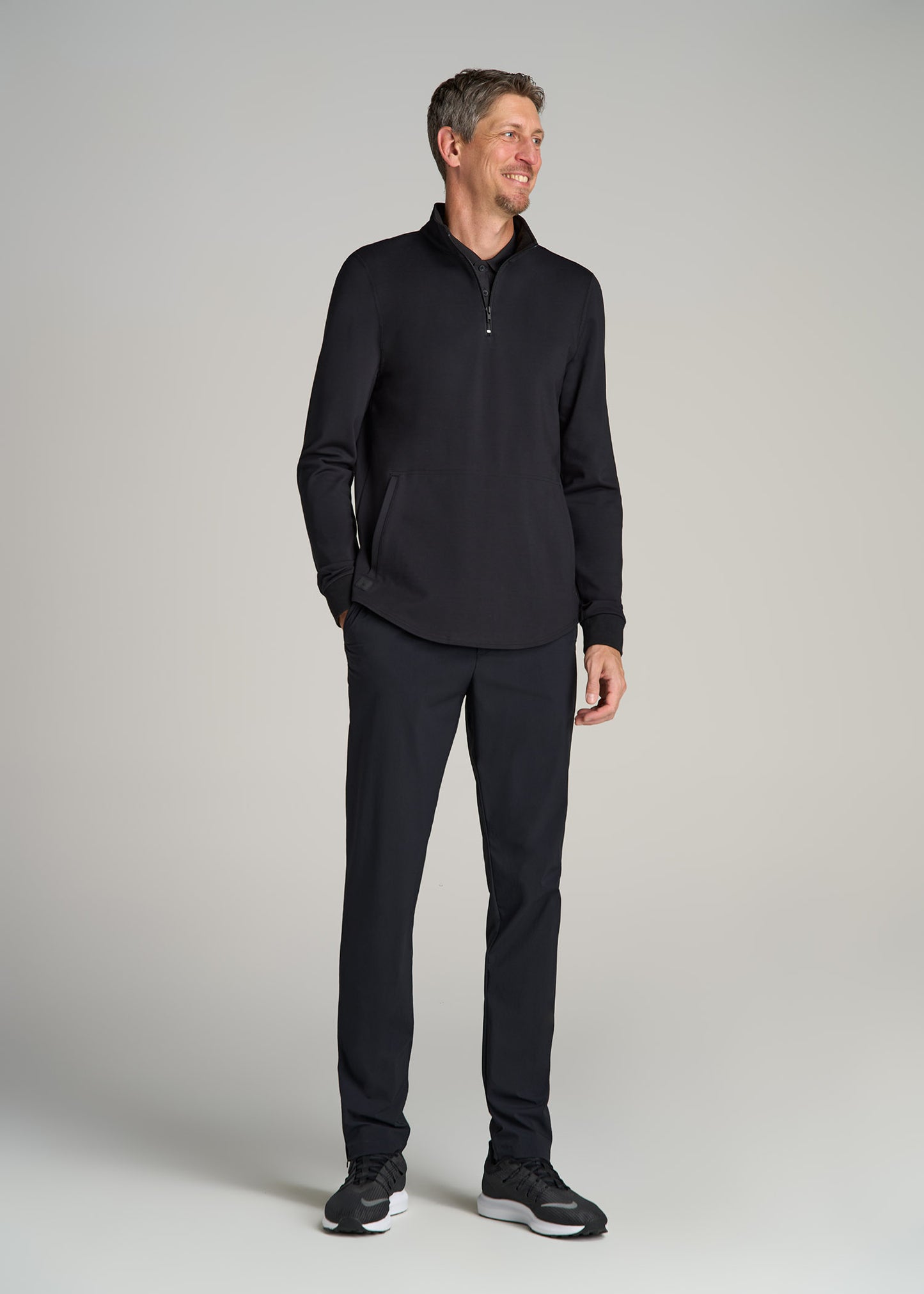 A tall man wearing American Tall's Fairway Popover Tall Men's Sweatshirt in Black and black pants.