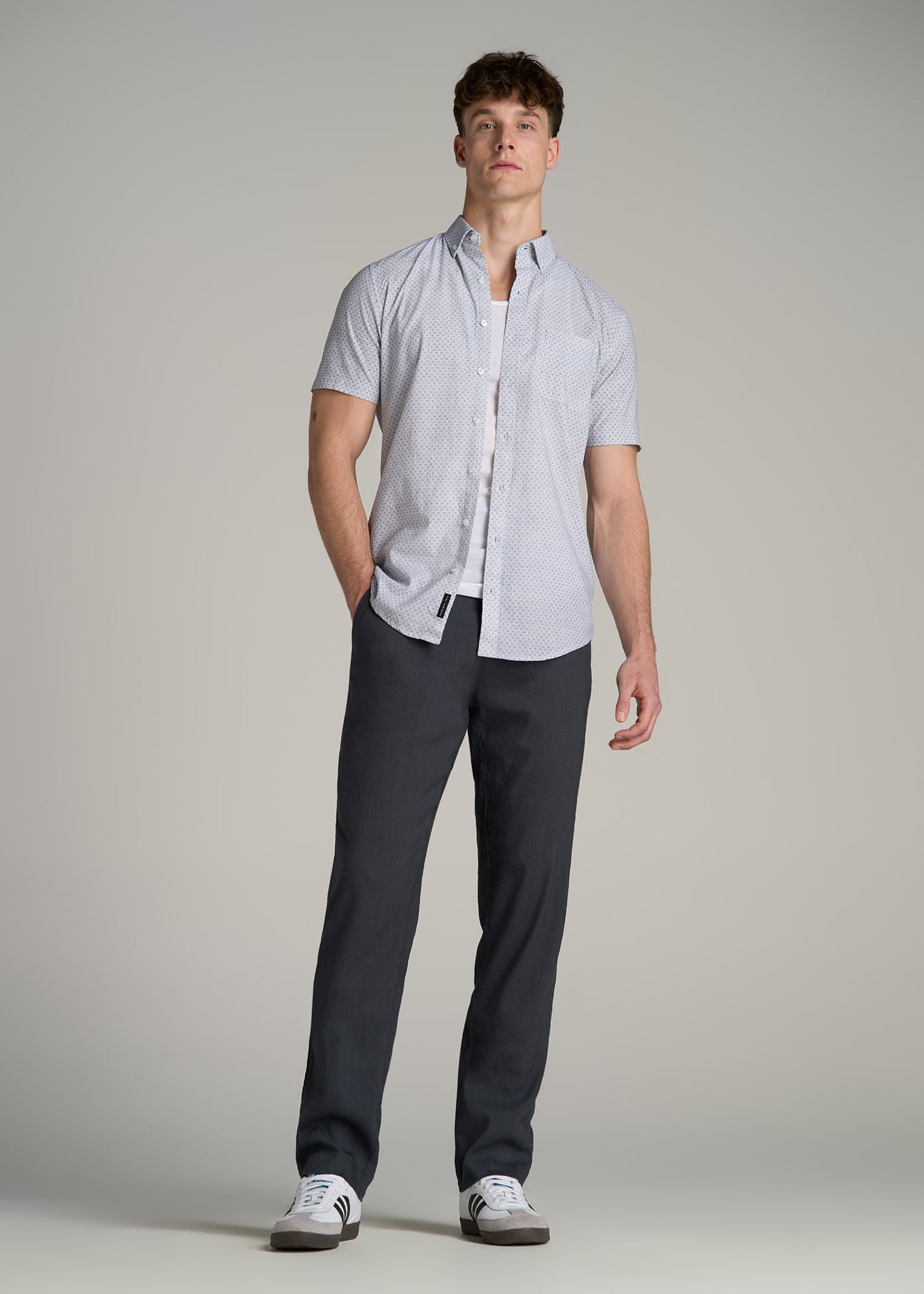 A tall man wearing American Tall's Short Sleeve Shirt for Tall Men in Navy Pindot, Men's Tall Ribbed Undershirt Tank Top in Bright White and Garment Dyed Linen Casual Pants for Tall Men in Heron Grey.