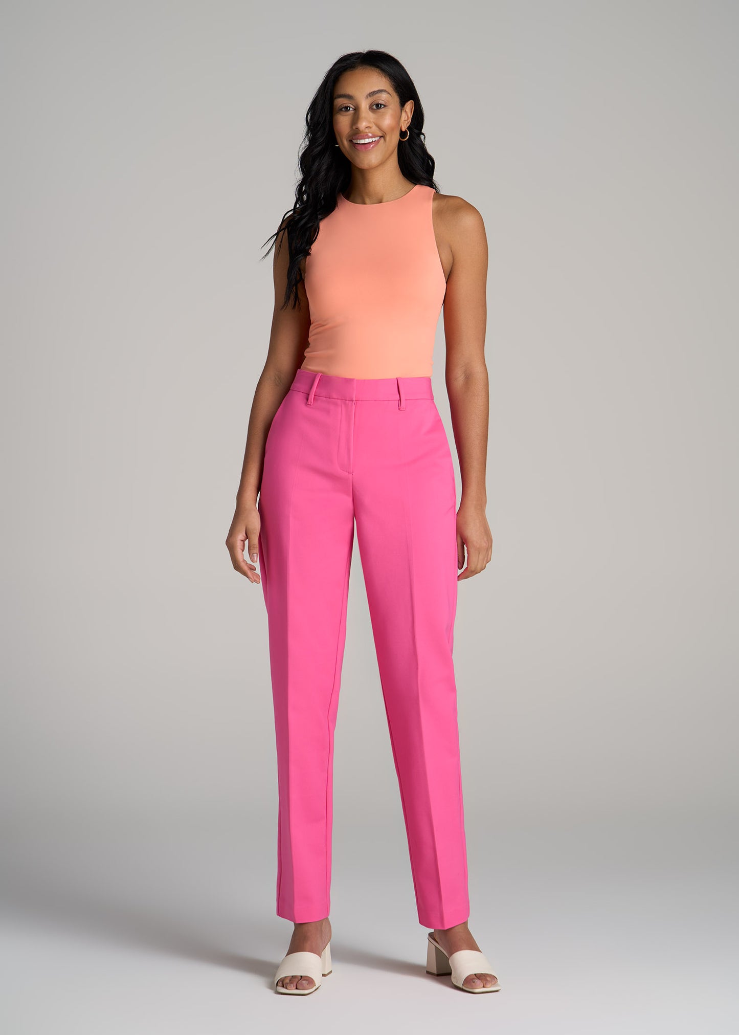 A tall woman wearing American Tall's Flat Front Tapered Dress Pants in Cosmo Pink and High Crewneck Sleeveless Bodysuit in Apricot Crush.